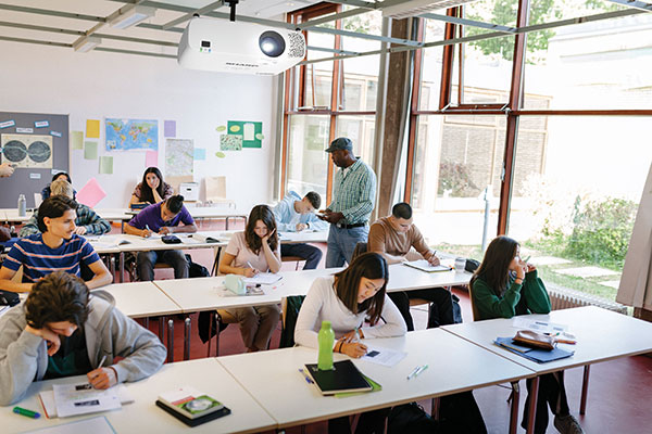 A young boy smiles in a classroom with a projector in the foreground and classmates seated behind him.